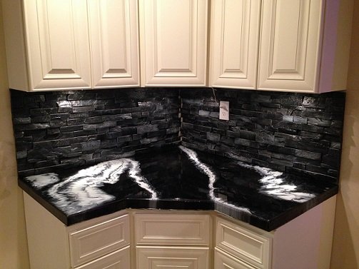 A kitchen backsplash of near-black stacked stone running around an inside corner below white cabinets, above a black and white marbled concrete countertop.