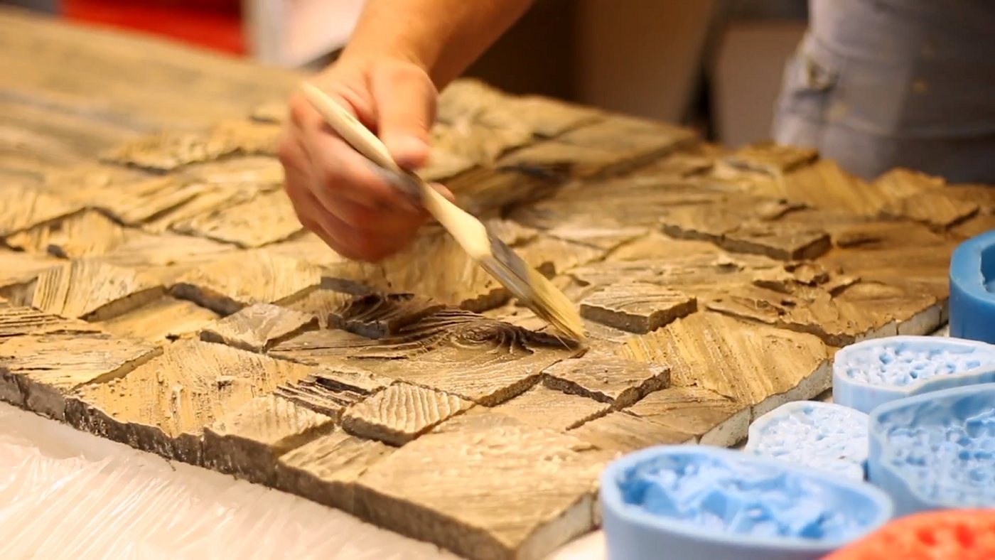 A hand brushing brown colour onto a cast concrete stone-pattern panel, with blue silicone molds in the foreground.