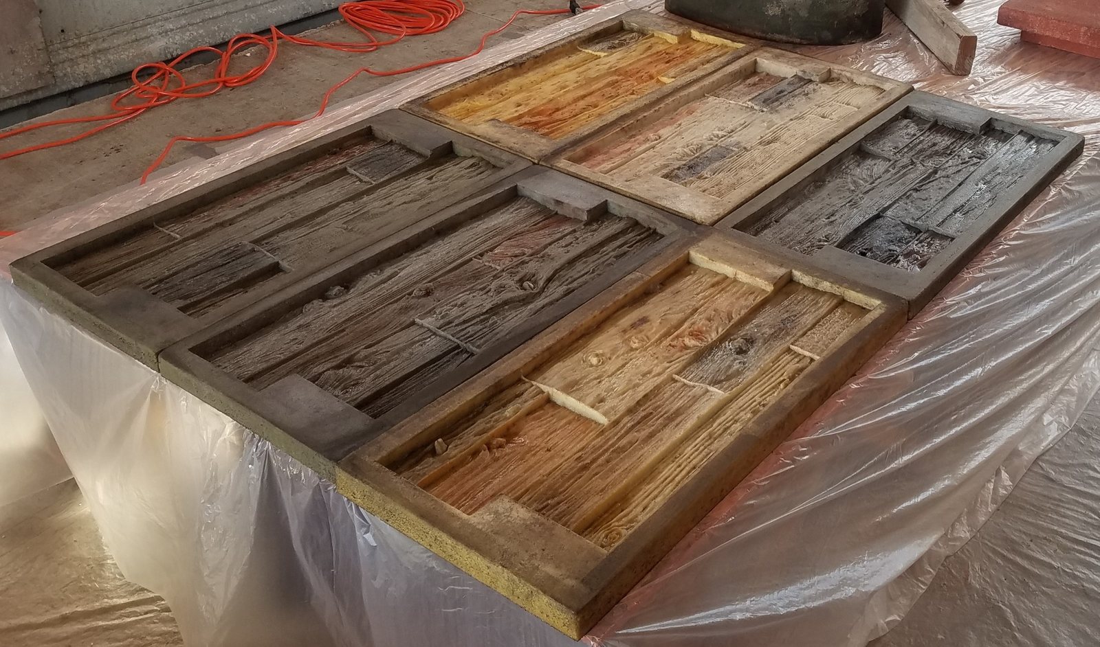 Several large rectangular concrete barn wood molds laid out on a plastic-covered bench in a workshop, showing plank grain and knots in different colours.