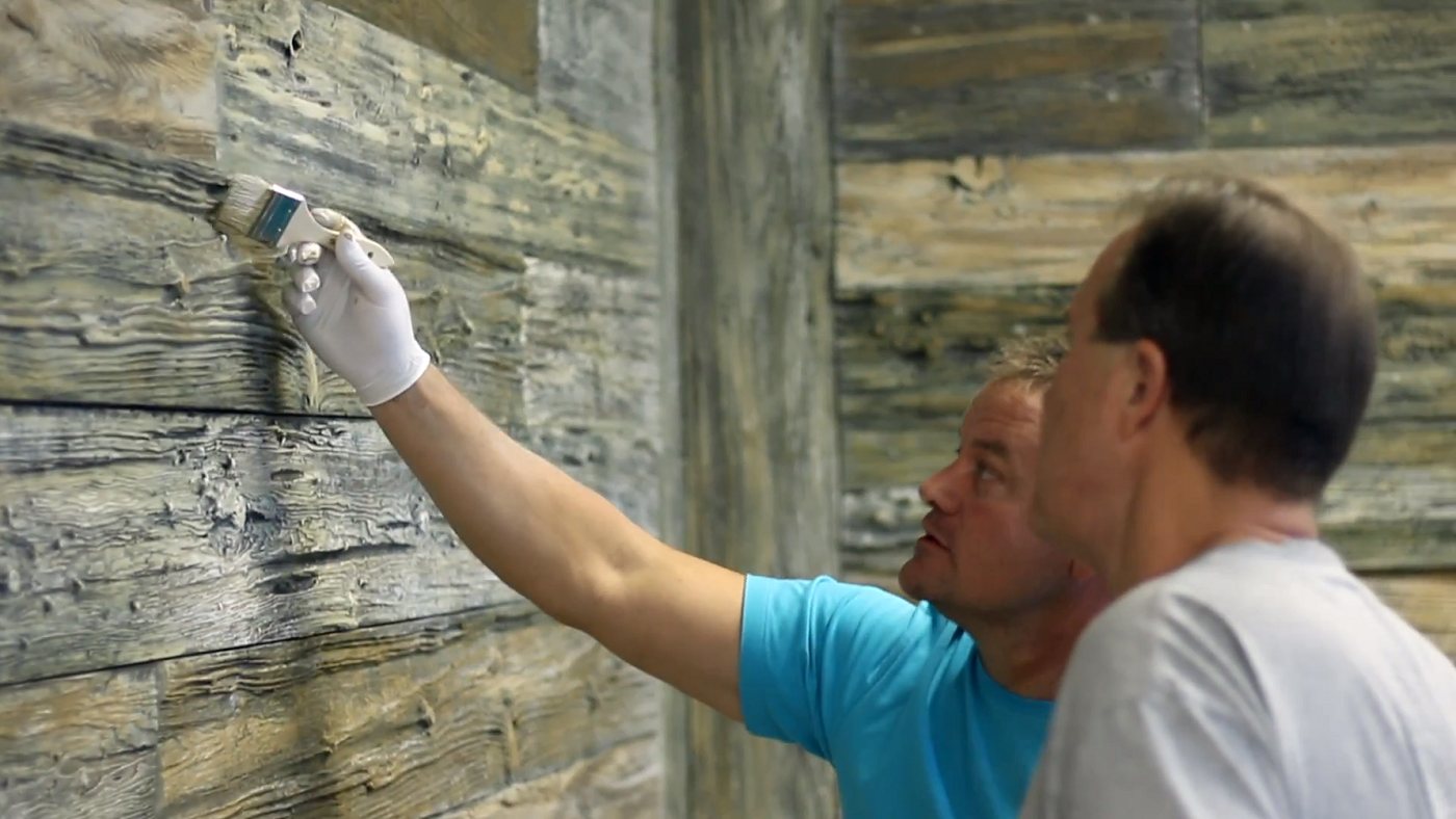 Two men staining a cast concrete barn wood wall with a brush, working across weathered grey and gold planks.