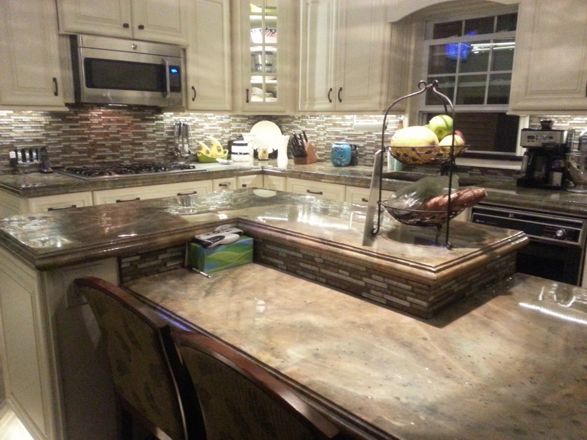 A kitchen with cream cabinets and glass tile backsplash, with concrete countertops coloured in mottled brown and grey across the perimeter, island and a raised breakfast bar.