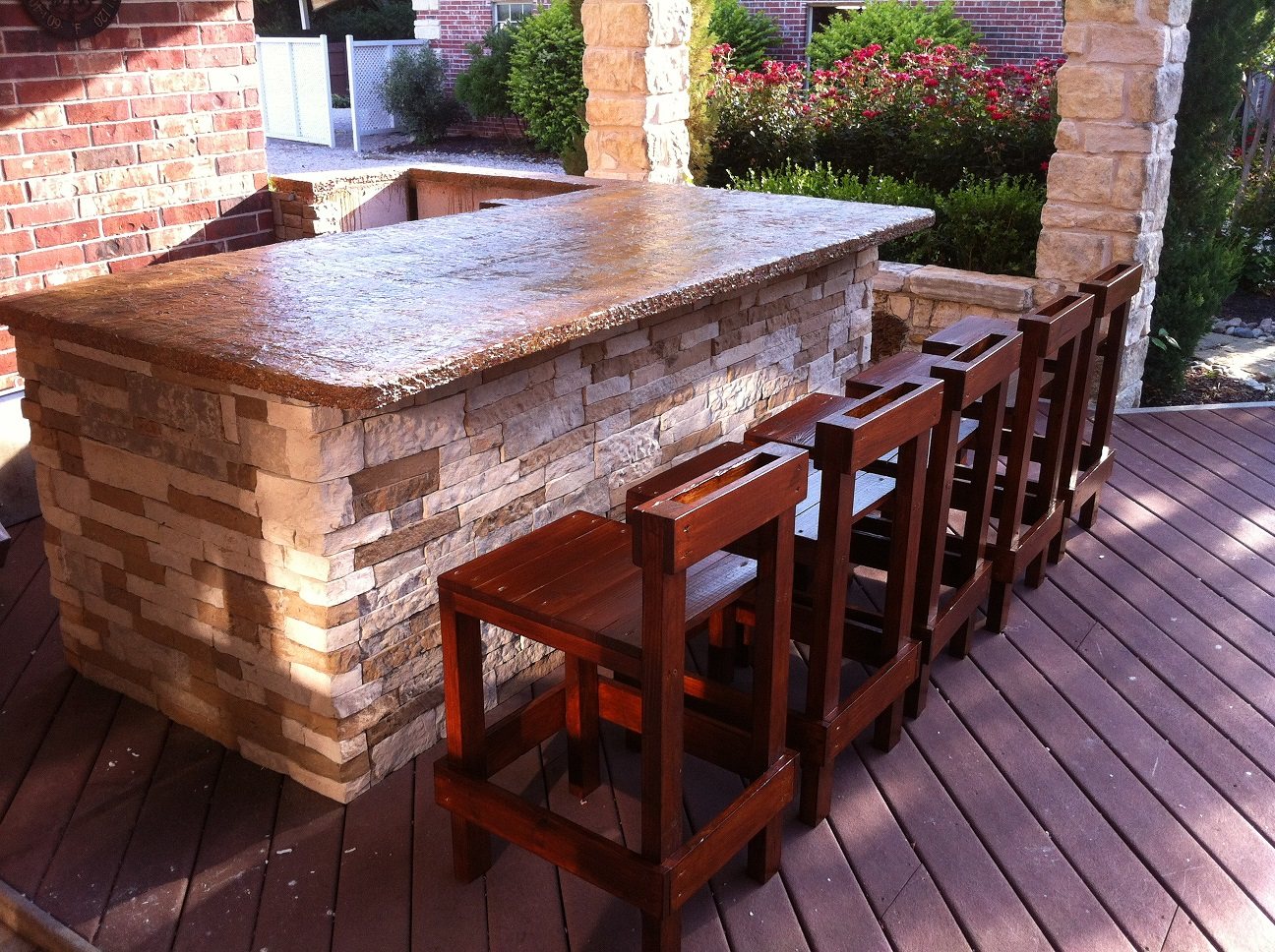 An outdoor L-shaped bar with a stacked stone base and a gold and tan concrete top, with wooden bar stools on a deck.