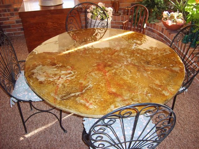 A round dining table with a concrete top coloured in amber, gold and rust with a glossy finish, surrounded by black wrought-iron chairs.