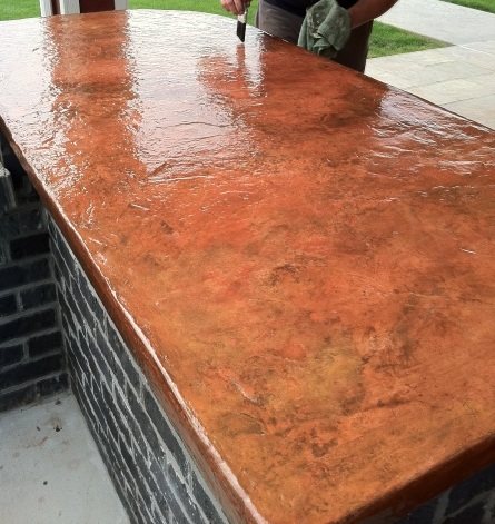 A person applying sealer to an outdoor concrete bar top coloured in copper and orange tones, the wet sealer bringing up a high shine.