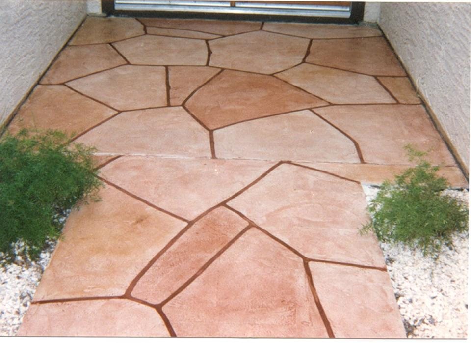 A small entry landing finished in terracotta concrete stamped as random flagstone with deep red grout lines, edged by white gravel and shrubs.