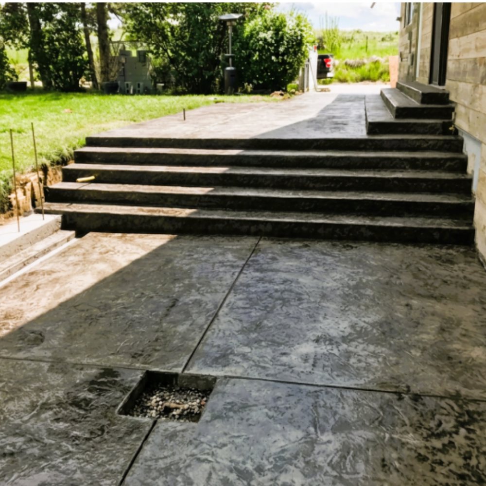 A charcoal grey textured concrete patio with two flights of matching steps leading up to a house, with green lawn and countryside behind.