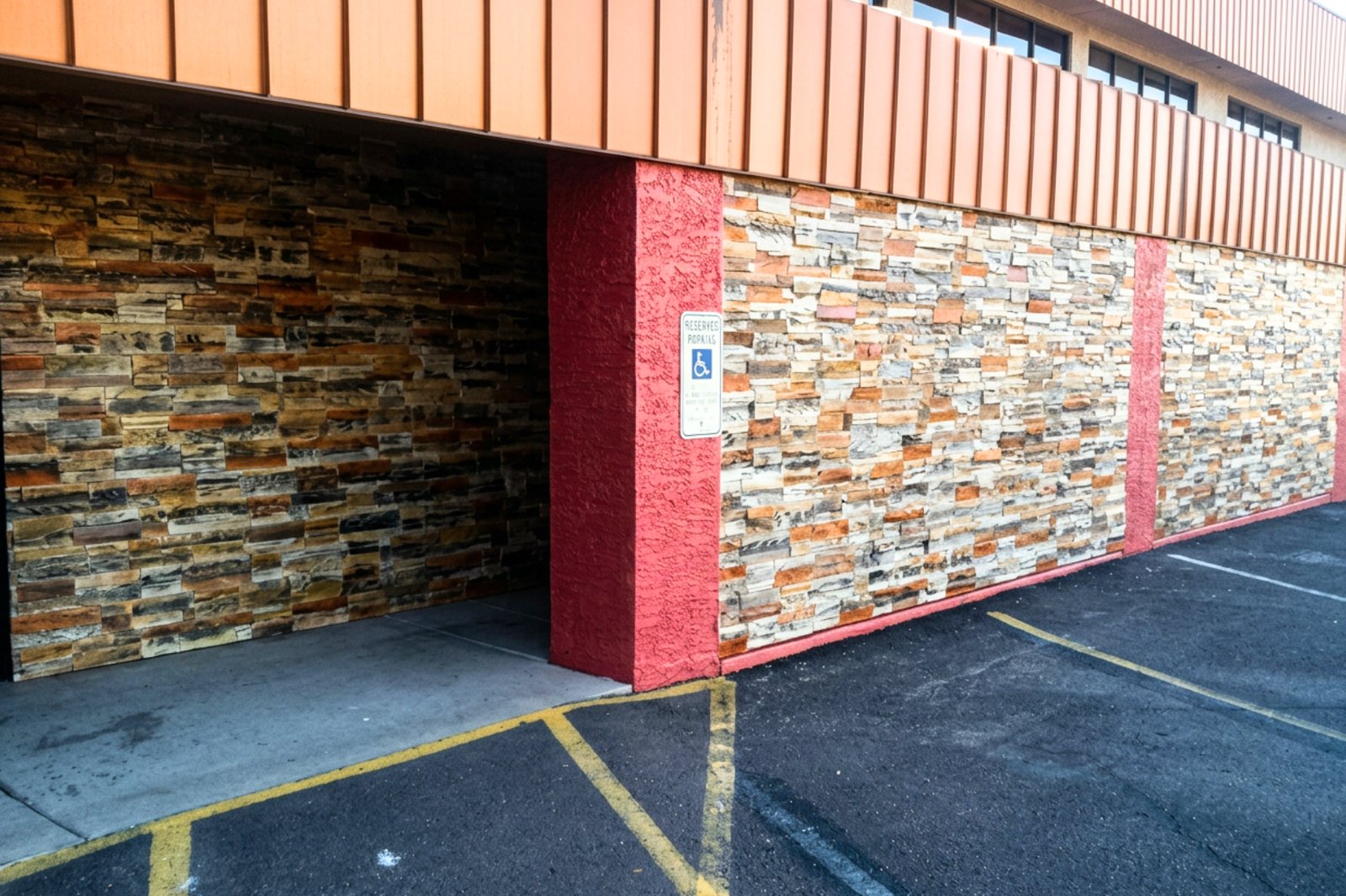The side of a commercial building clad in concrete stacked stone panels in two different colourways, separated by red-painted columns, beside a car park.