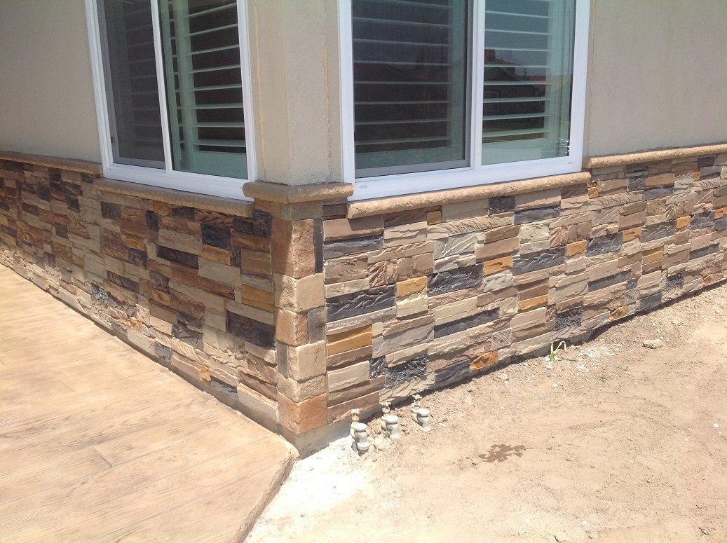 The corner of a house below a bay window, clad in tan, grey and dark brown stacked stone panels with a cast concrete sill, beside a stamped concrete patio.