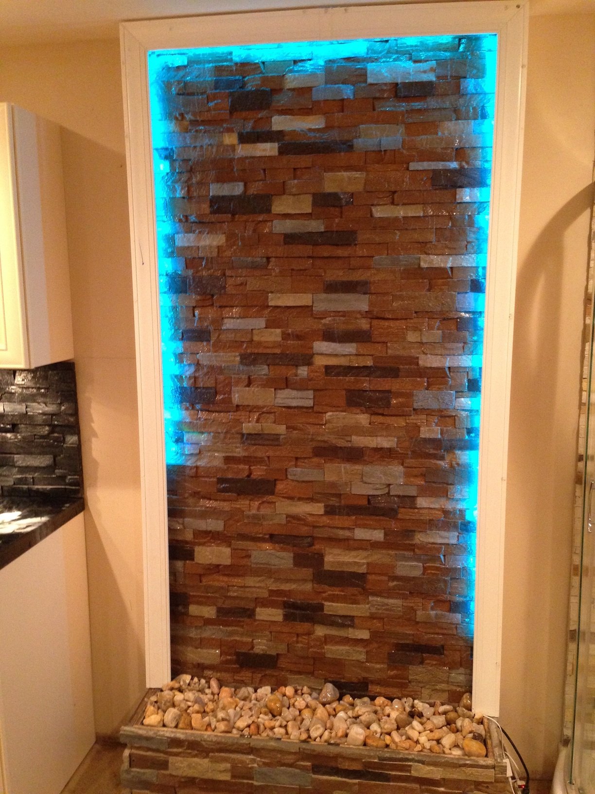 A tall recessed water wall feature clad in multi-coloured stacked stone panels with water running down the face, lit with blue LED light, over a bed of river pebbles.