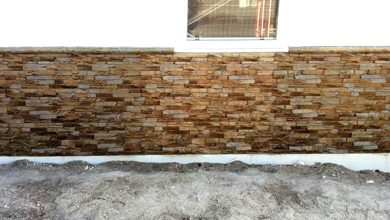 A long exterior wainscot clad in gold and tan stacked stone panels with a cast coping, on a new build with bare soil at its base.