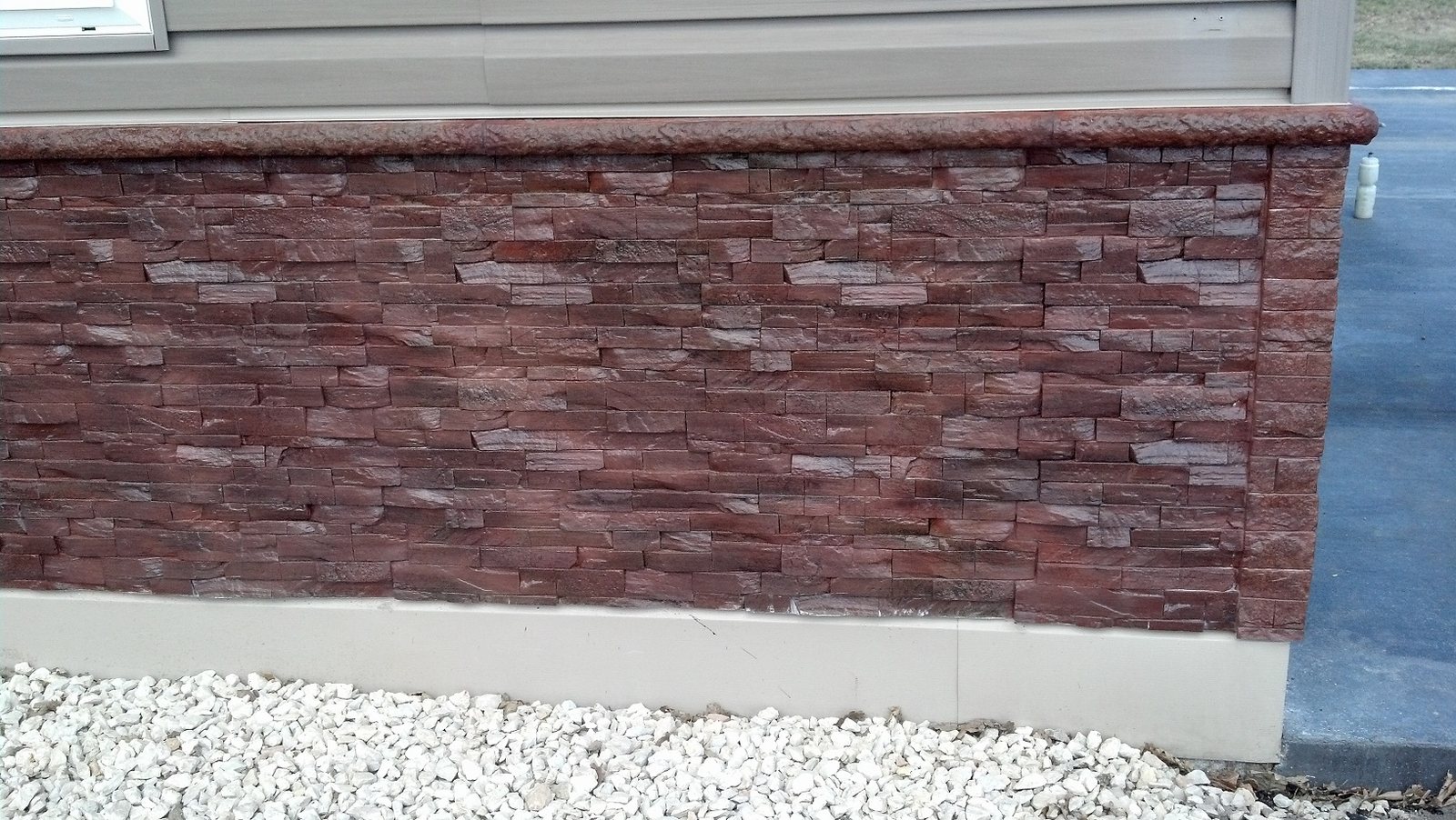 A house wainscot clad in deep red and maroon stacked stone panels with a matching cast cap, against grey siding above white gravel.