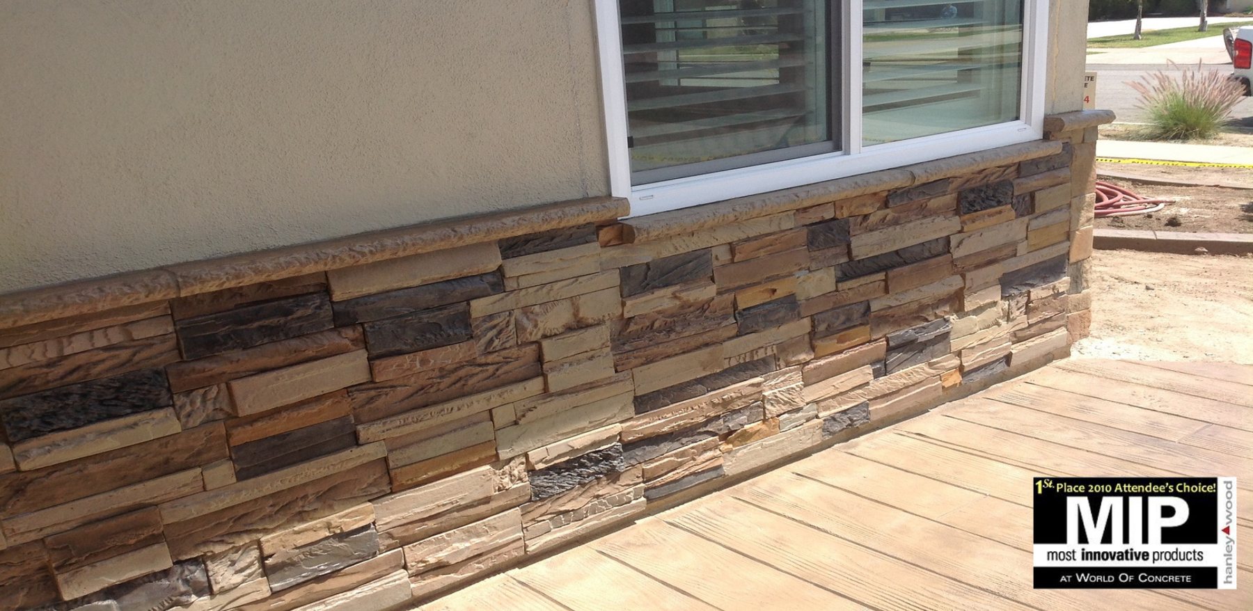 An exterior wall below a window clad in warm brown and gold stacked stone panels with a cast sill, above a concrete deck finished to look like timber planks.
