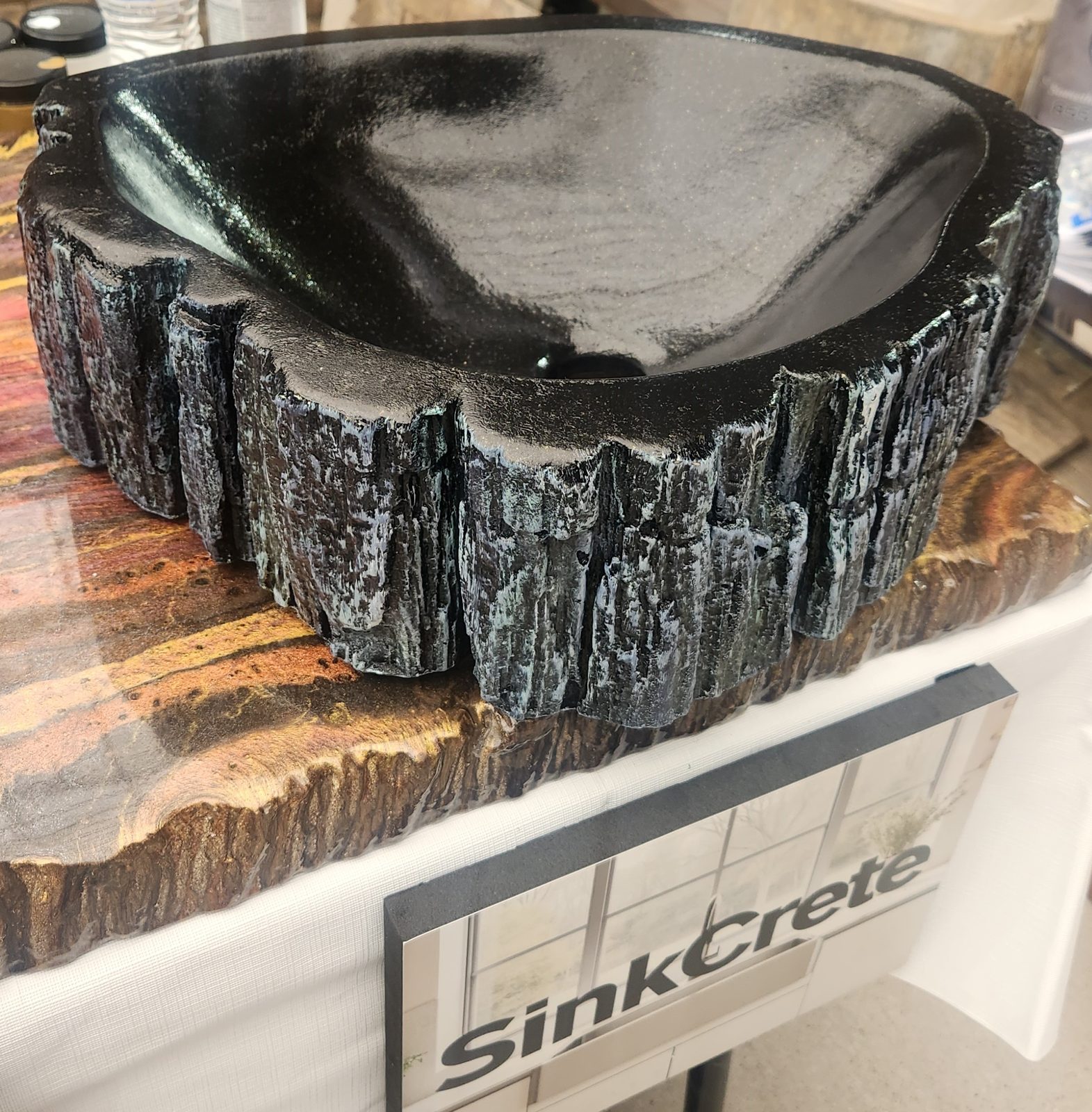 A black concrete vessel sink with silvered bark texture on a trade-show stand marked SinkCrete.